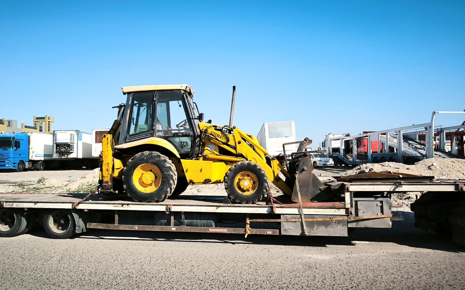 A yellow construction vehicle is seen on a flatbed trailer in an industrial area under a clear blue sky.